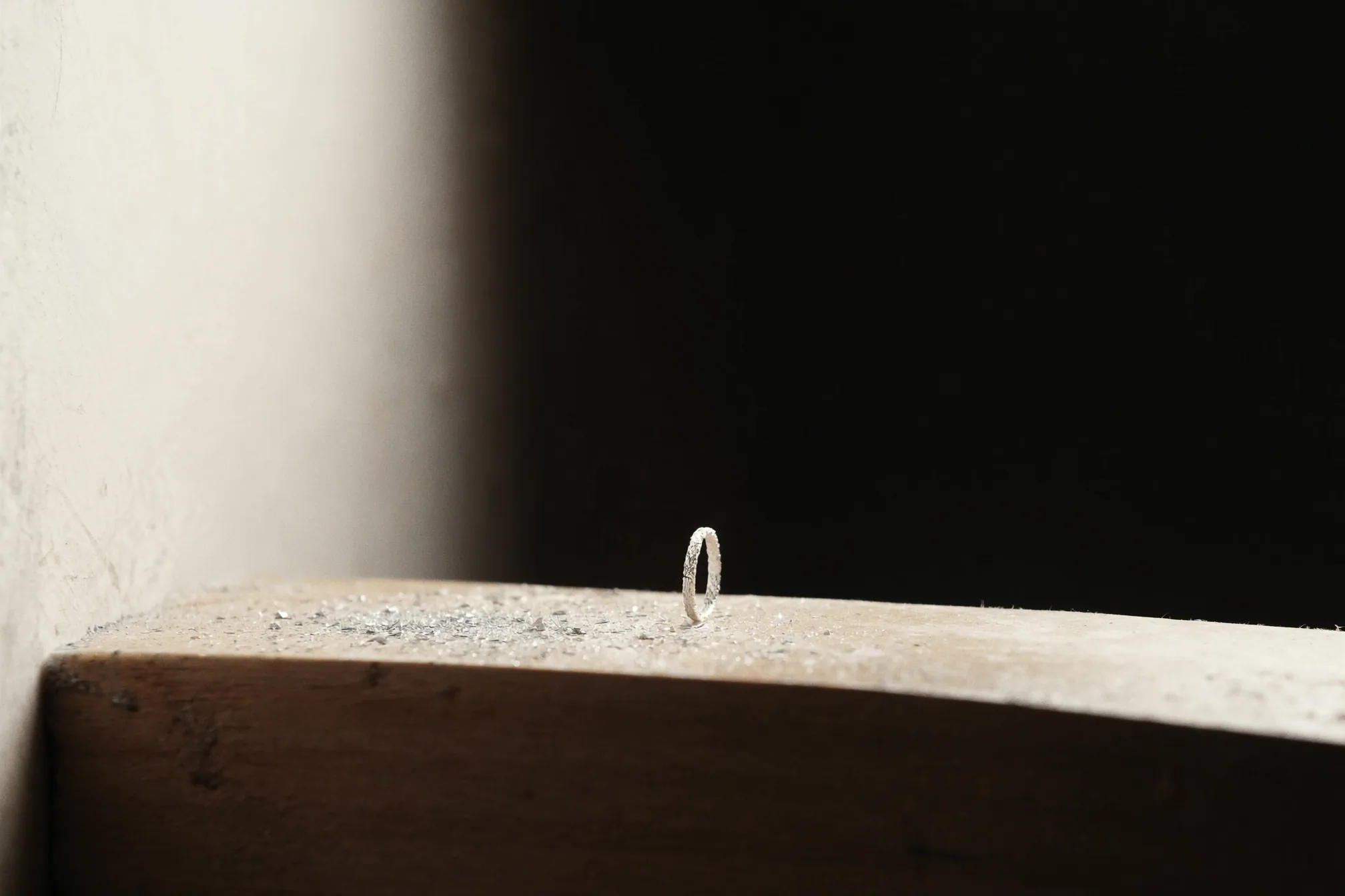 A silver ring standing upright on the edge of a wooden bench dusted with filings, a dark room behind it.