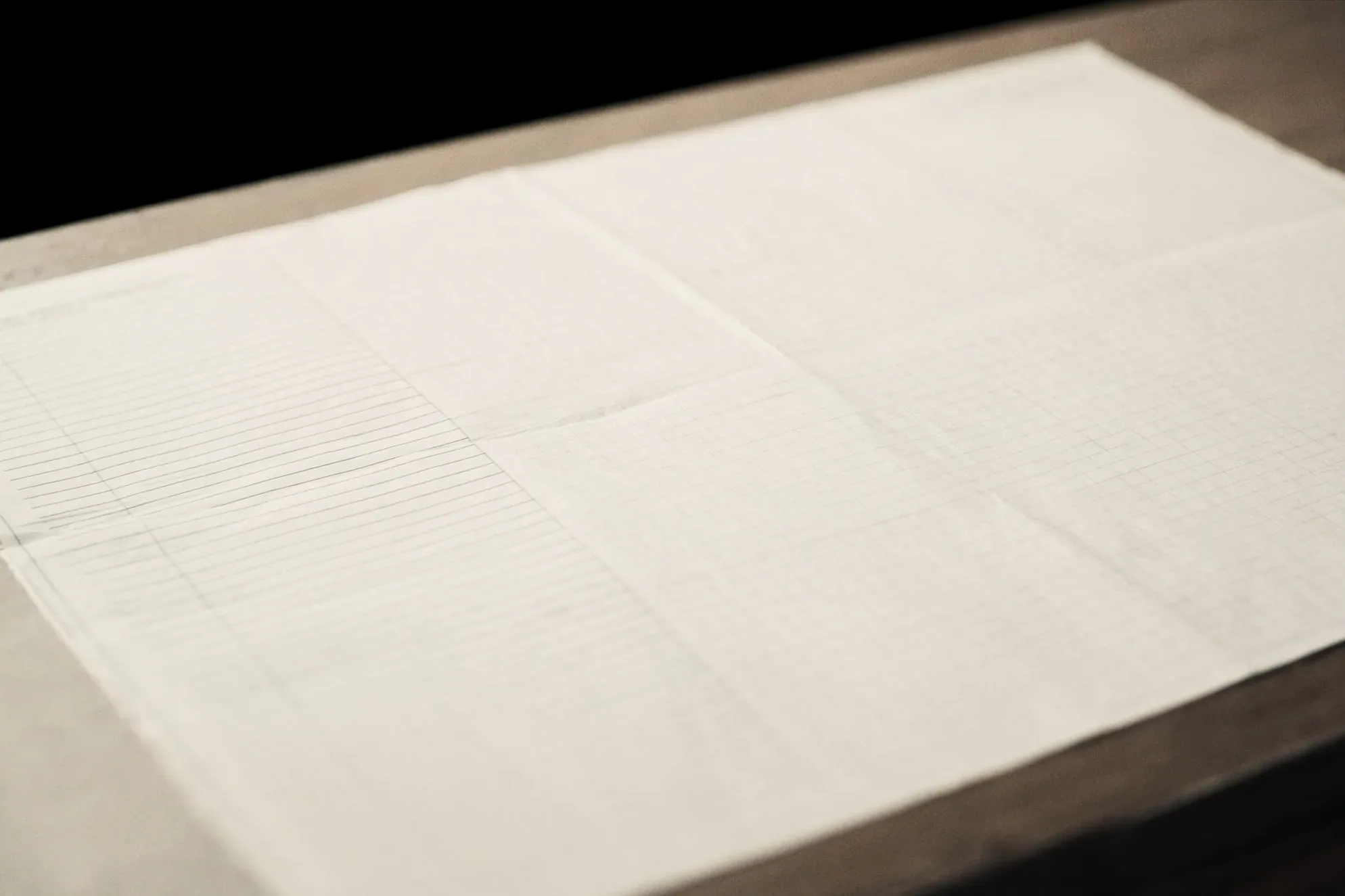 A wide sheet of ruled and gridded paper lying flat on a dark table, its folds catching light from the right.