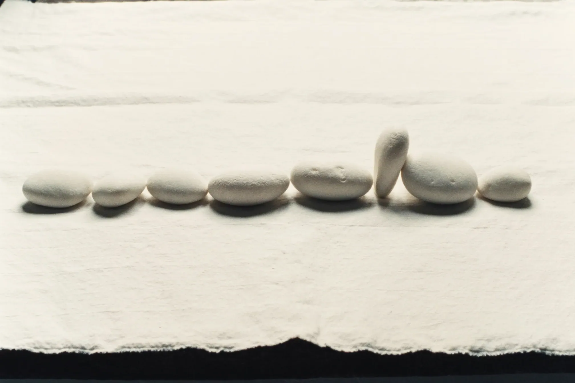Eight pale stones sit in a row on white cloth, one standing upright against the others, dark surface below.