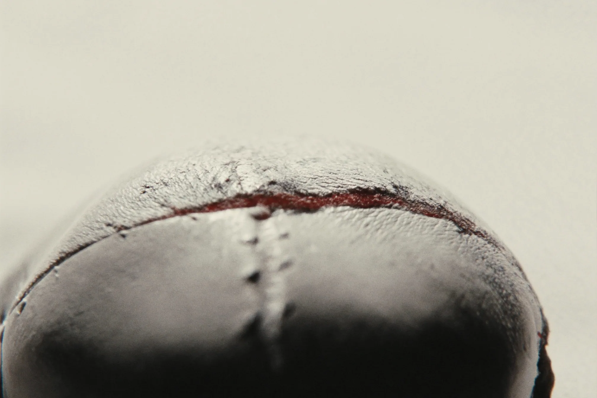Close view of the top of a worn leather ball, its stitched seam and dark red edge against a pale ground.